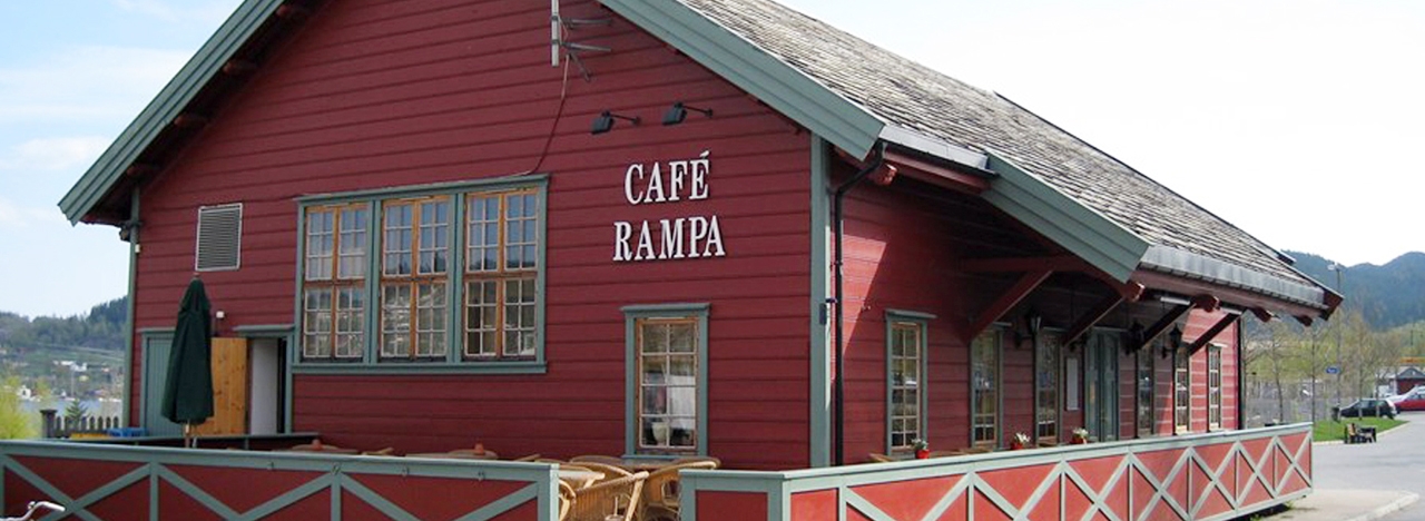 Det røde pakkhuset som huser Cafè Rampa, med hvitt skilt på veggen og grønt rekkverk rundt terrassen